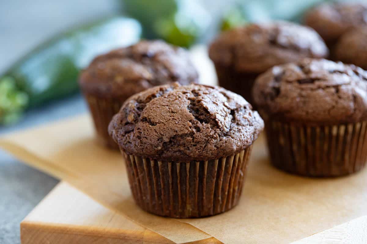 Chocolate Zucchini Muffins on a wooden board with fresh zucchini behind.