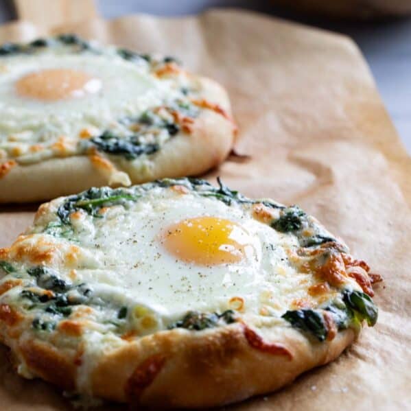 Two individual sized pizzas topped with creamed spinach and an egg on parchment paper.