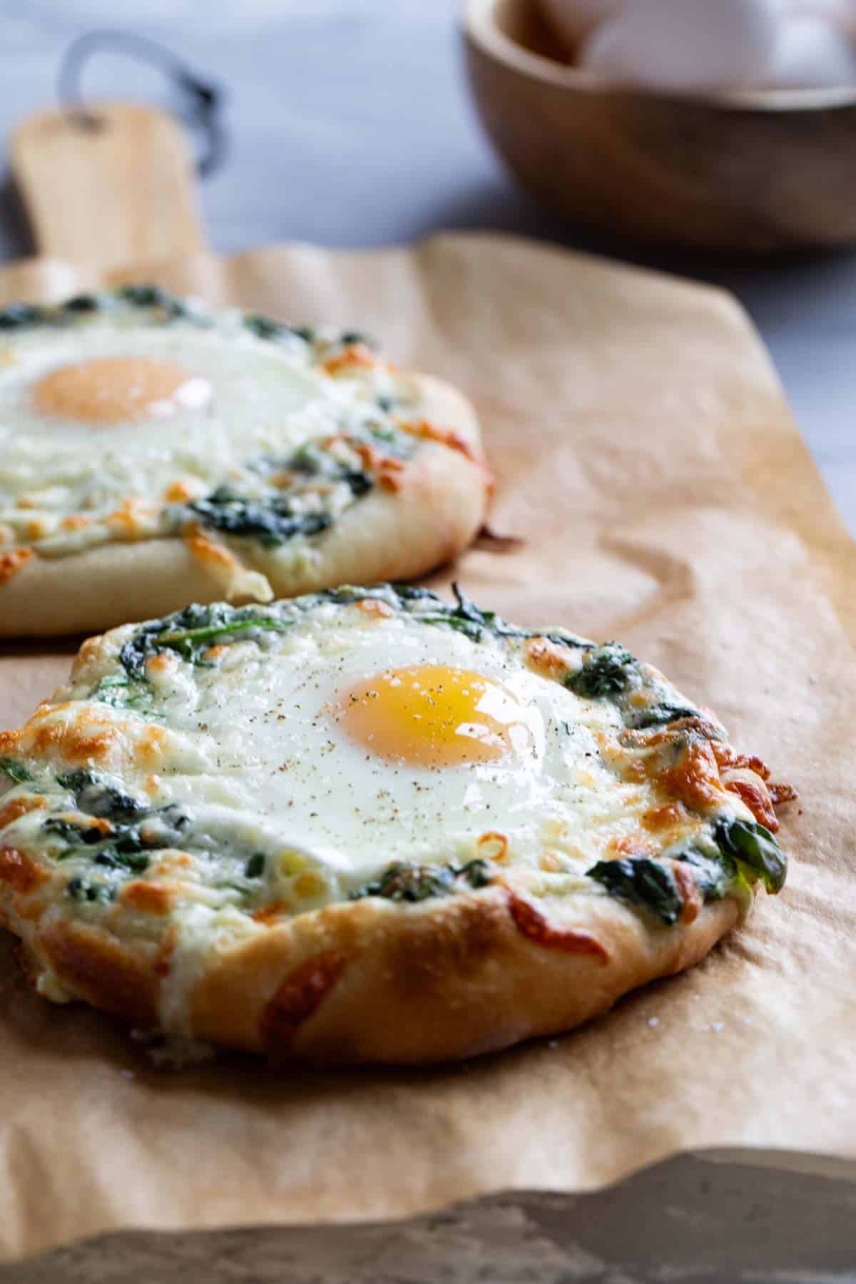 Two individual sized pizzas topped with creamed spinach and an egg on parchment paper.