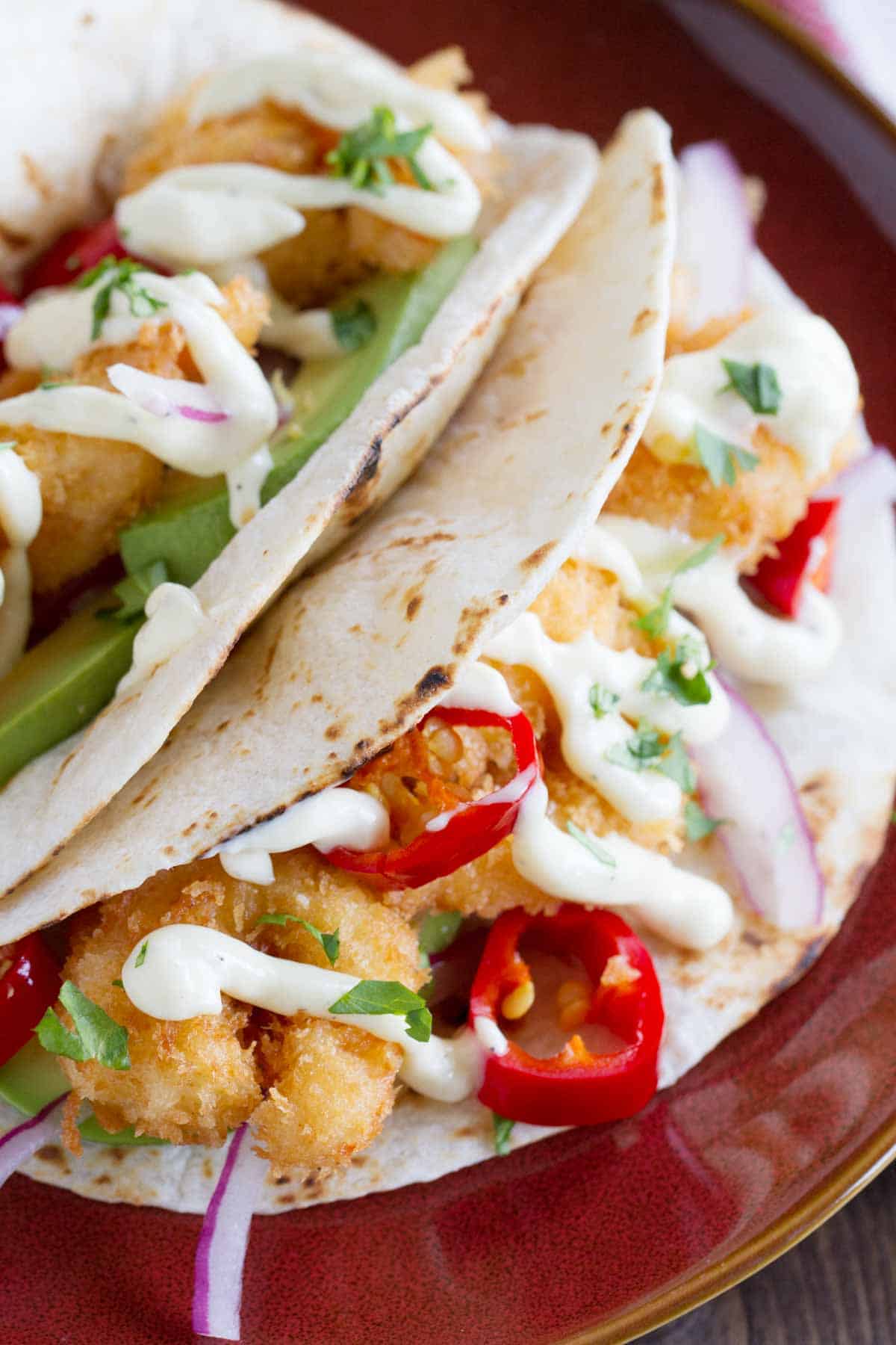 Two crispy shrimp tacos on a plate, topped with red onions, peppers, and homemade mayonnaise.