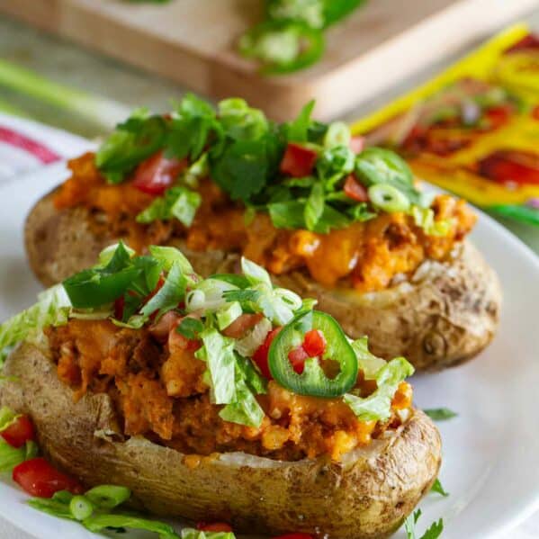 Two taco stuffed potatoes on a plate, topped with lettuce, tomatoes, and jalapeños.