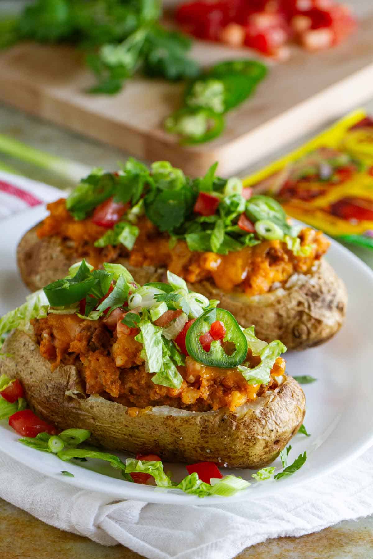 Two taco stuffed potatoes on a plate, topped with lettuce, tomatoes, and jalapeños.