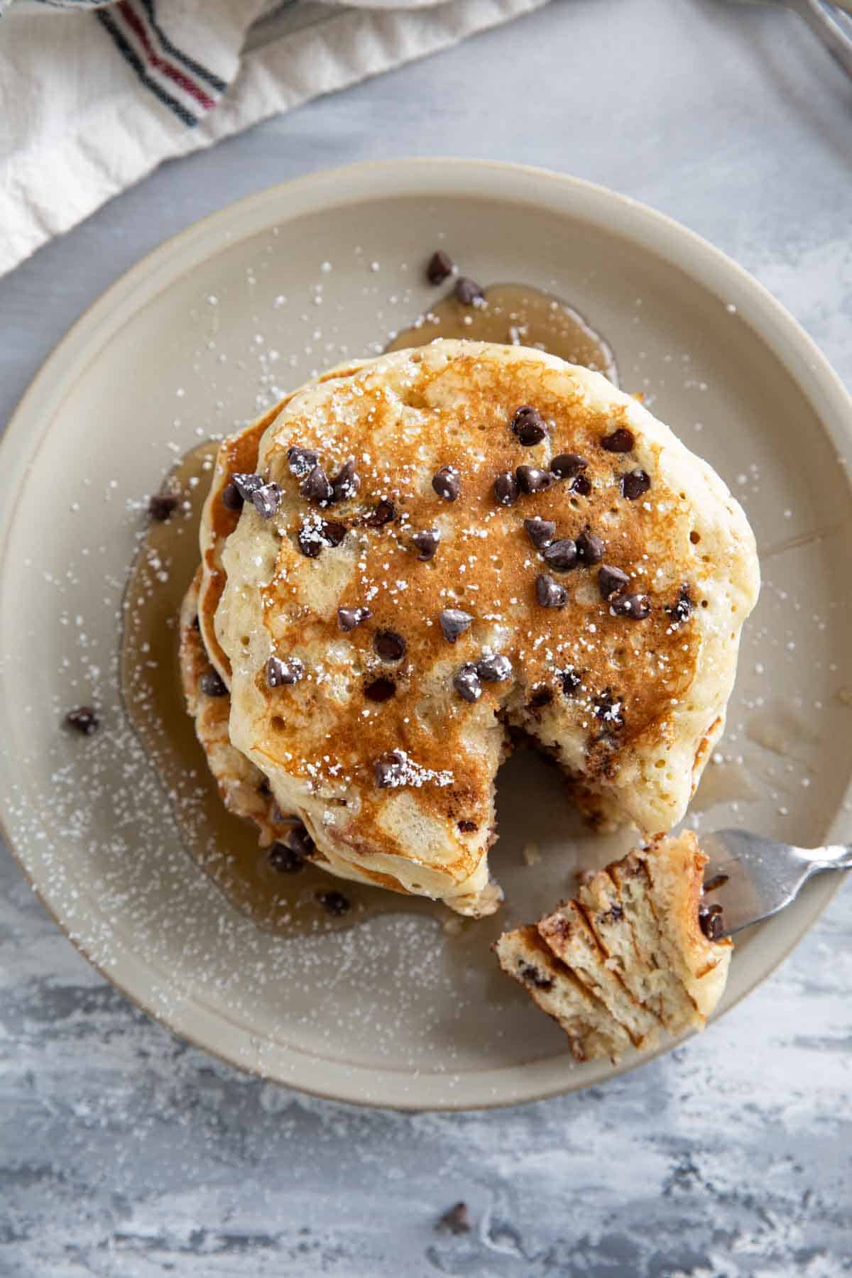 Stack of chocolate chip pancakes topped with syrup and powdered sugar.