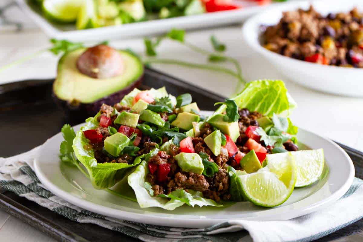 Taco lettuce wraps on a white plate with more filling behind them.