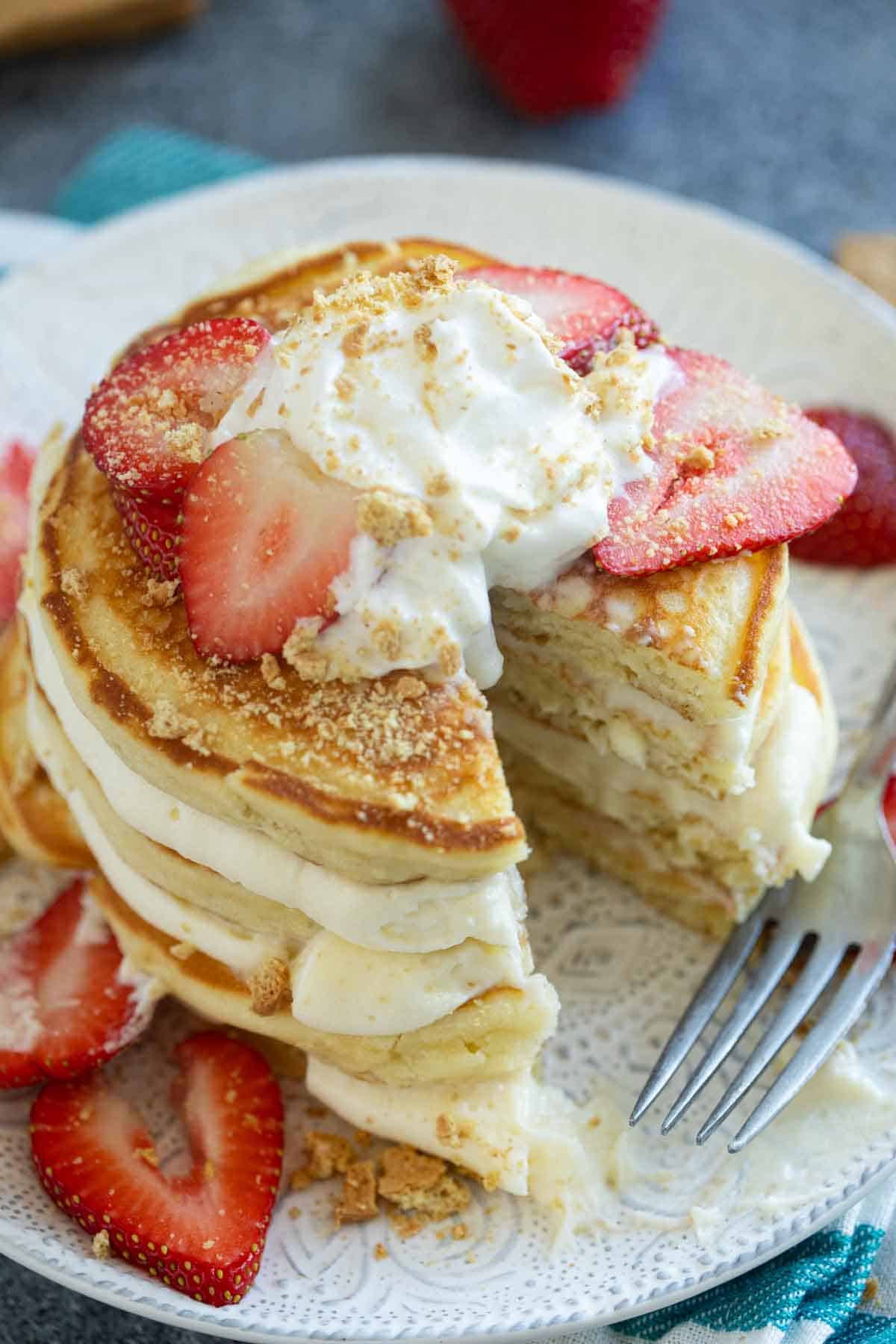 Stack of Cheesecake Pancakes topped with whipped cream, strawberries, and graham cracker crumbs.