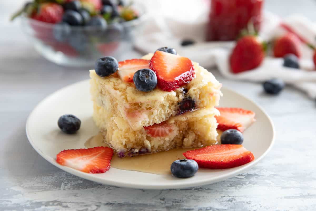 Two slices of sheet pan pancakes on top of each other, with syrup, strawberry slices, and blueberries.