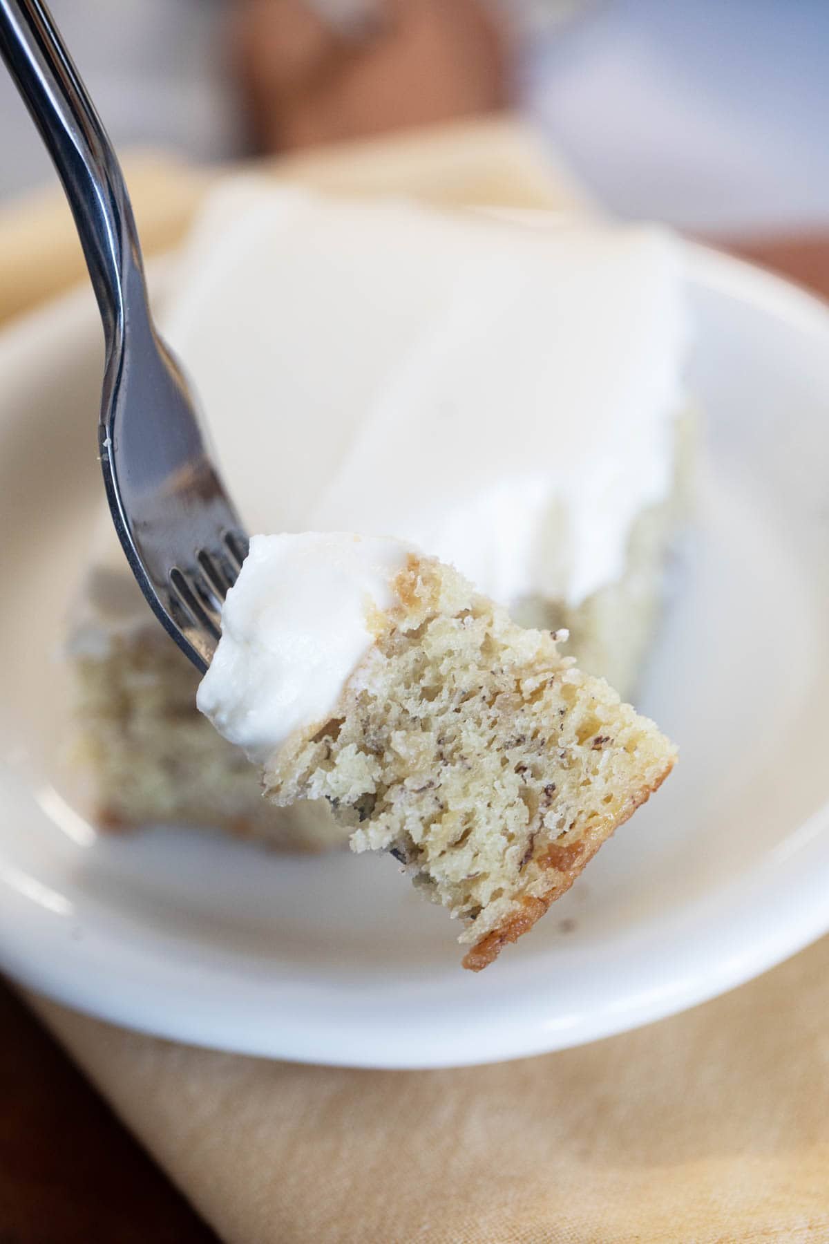 Bite of banana cake with cream cheese frosting on a fork in front of a slice of cake.