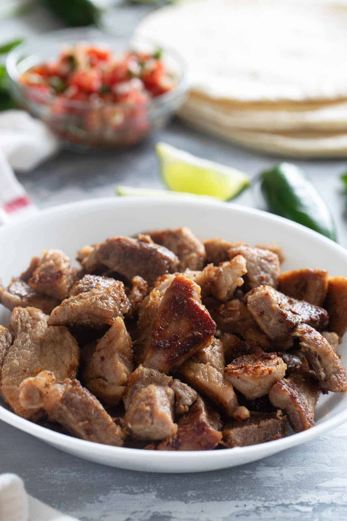 Pork Carnitas in a bowl with tortillas, limes, and salsa in the background.