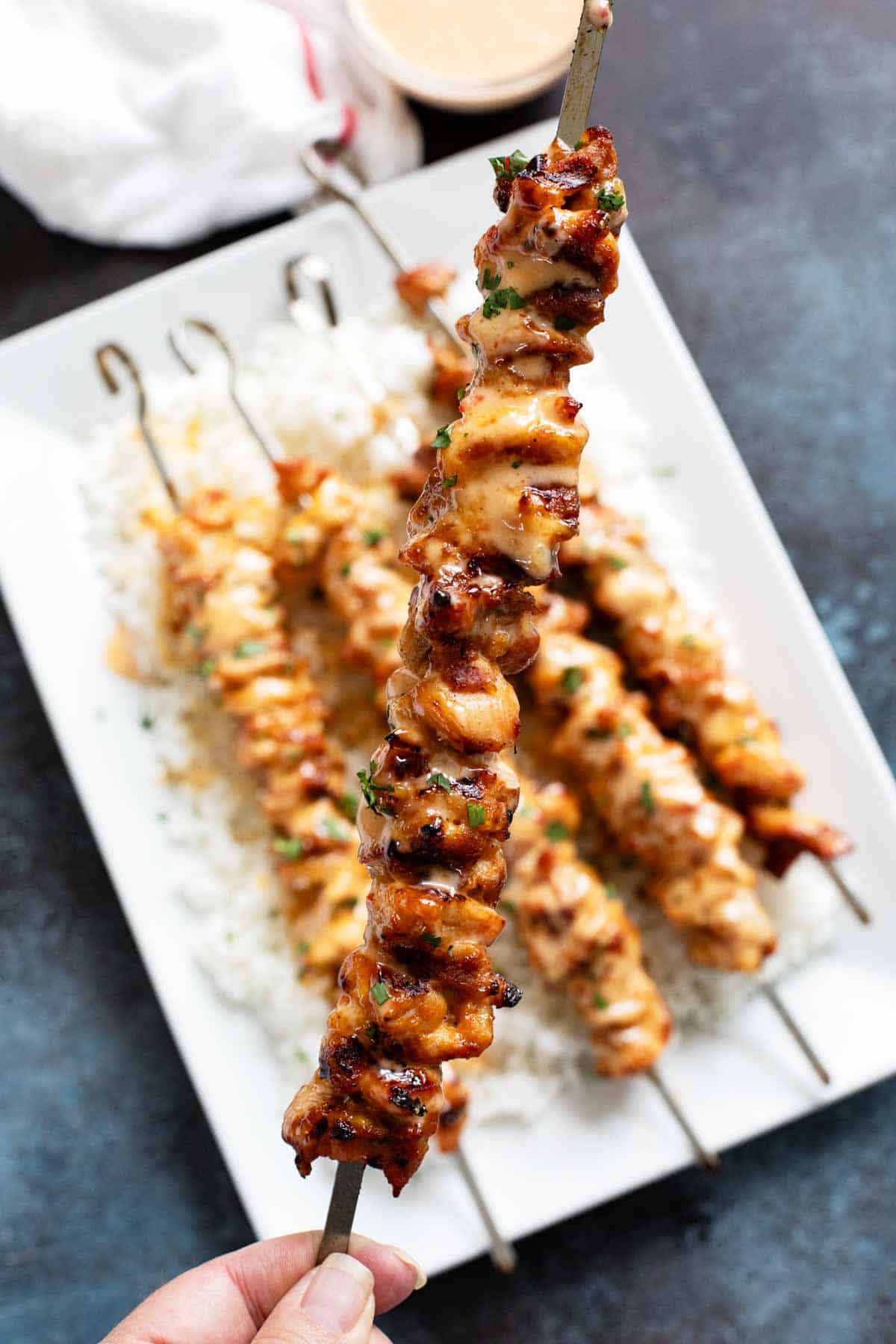 Holding up a bang bang chicken skewer with more on a plate below.