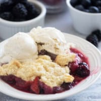 Berry Cobbler in a small bowl, topped with vanilla ice cream with fresh berries in the background.