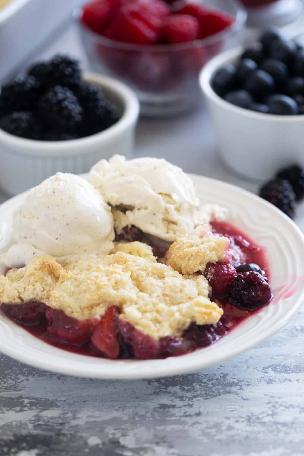 Berry Cobbler in a small bowl, topped with vanilla ice cream with fresh berries in the background.