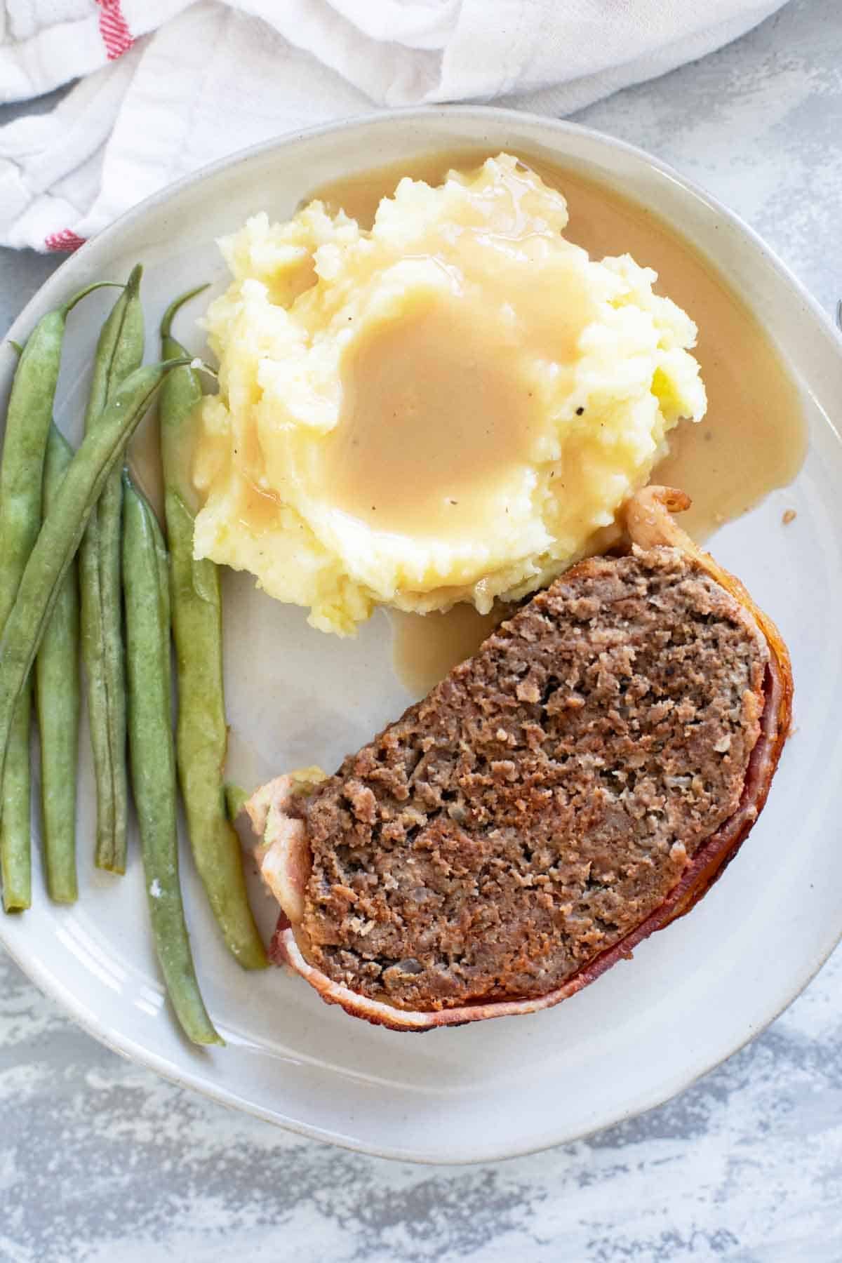 Plate with a slice of bacon wrapped meatloaf, mashed potatoes and gravy, and green beans.