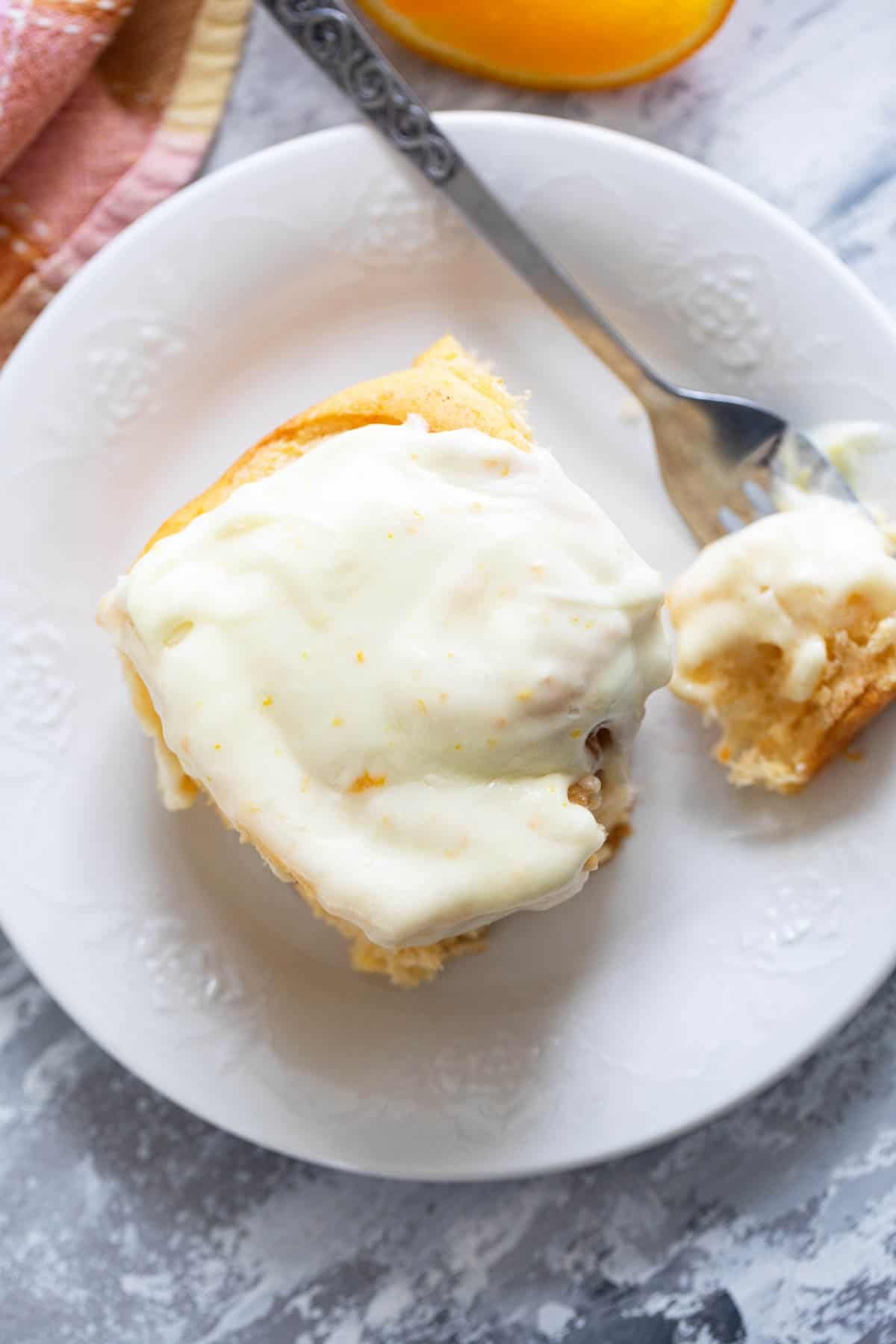 Sweet Potato Cinnamon Roll on a plate, topped with orange cream cheese frosting, with a bite taken with a fork.
