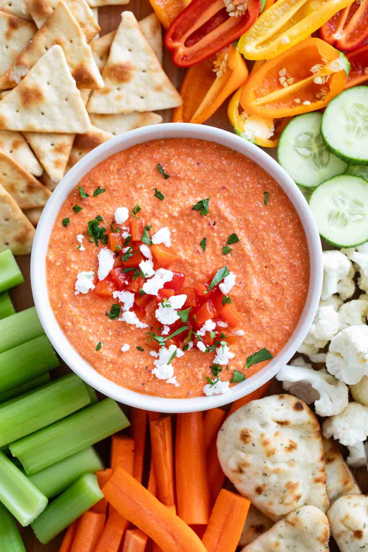 Bowl of Feta and Roasted Red Pepper Dip surrounded by vegetables, crackers, and naan.