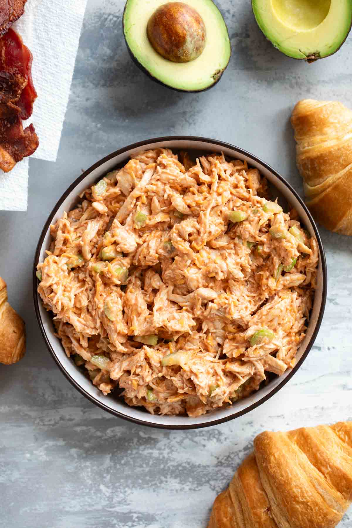 Bowl of BBQ Ranch Chicken Salad surrounded by croissants, avocado, and bacon.