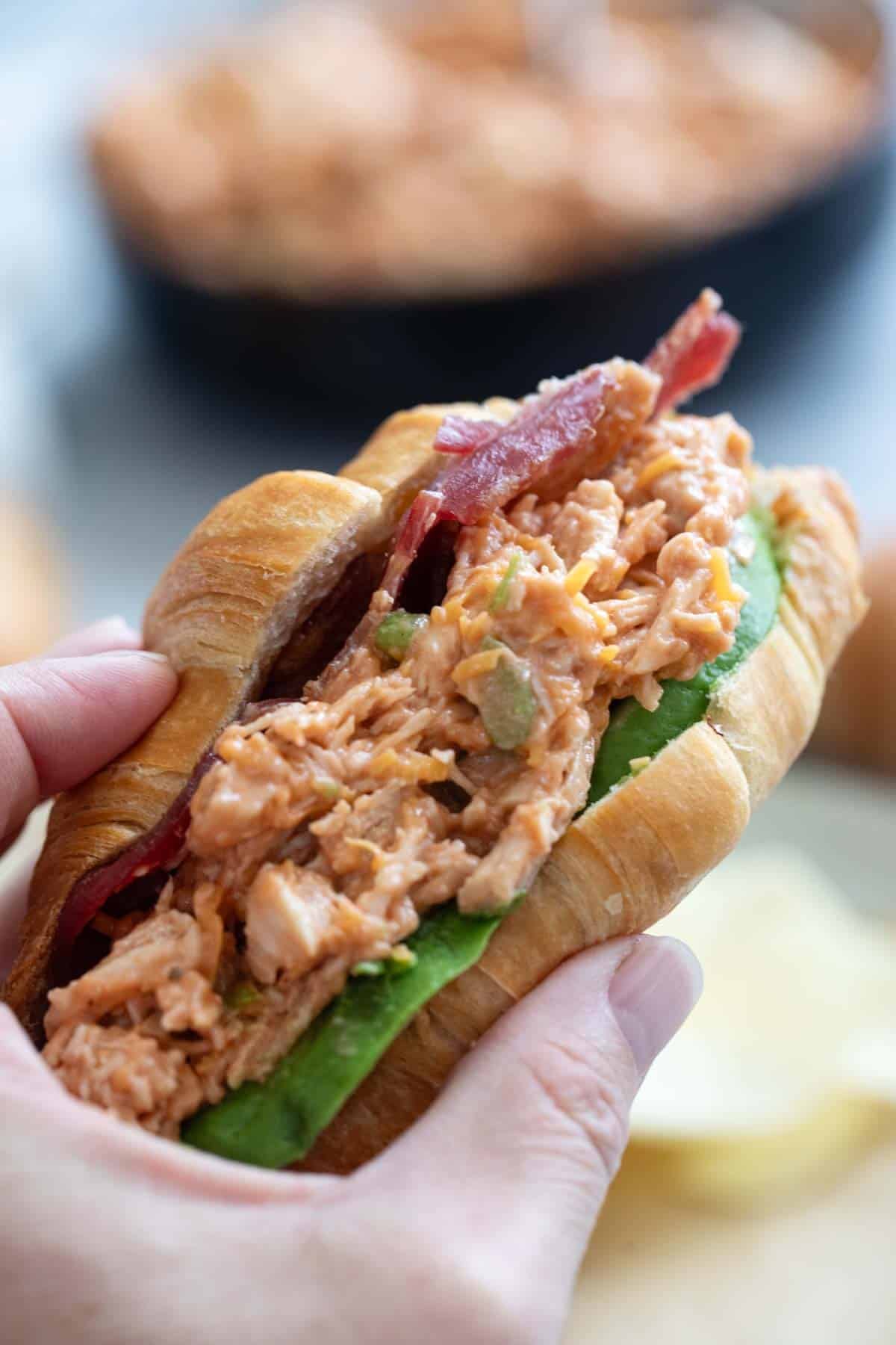 Hand holding a BBQ Ranch Chicken Salad Sandwich with bacon and avocado.