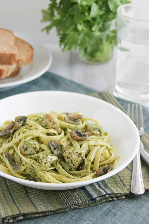 Serving of Chicken and Pasta with Cilantro Pesto in a white bowl, with bread and cilantro behind.