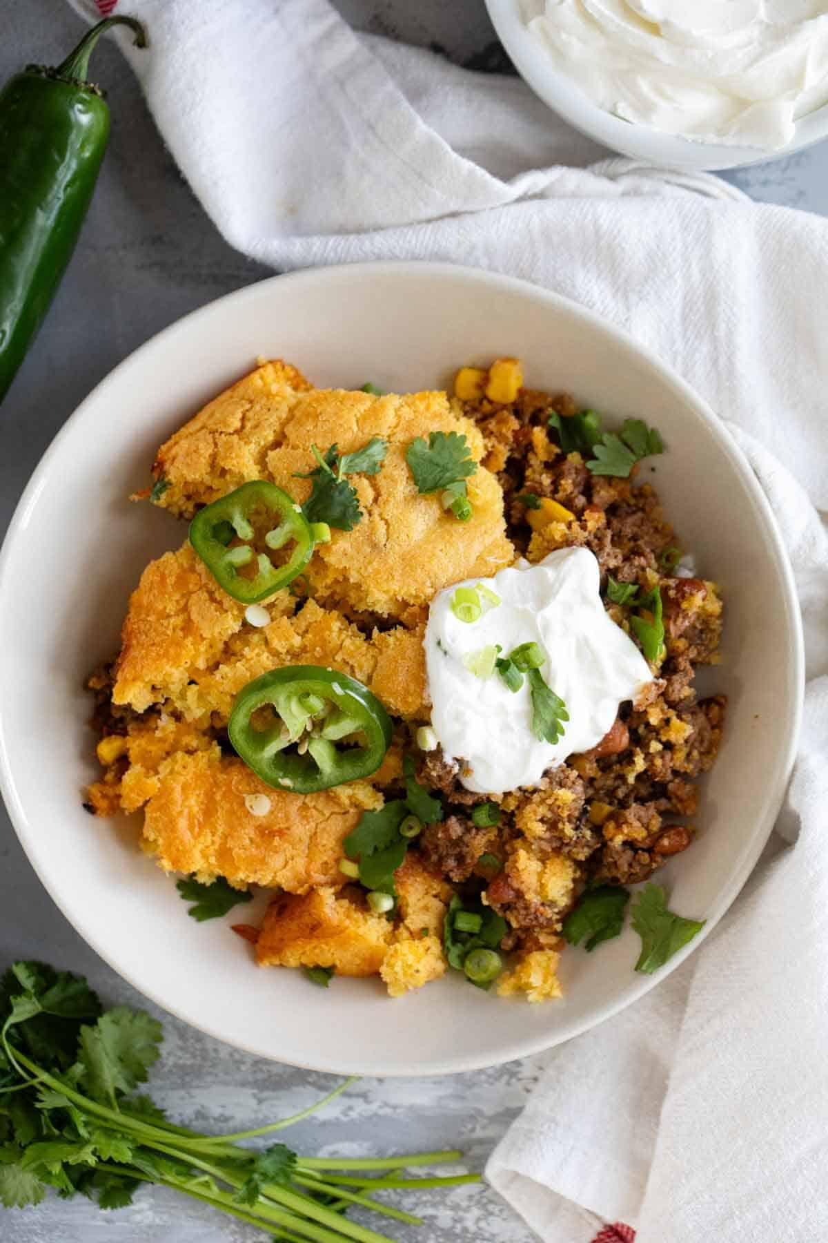 Shallow bowl with a serving of Chili Cornbread Casserole topped with sour cream, cilantro, and jalapeños.