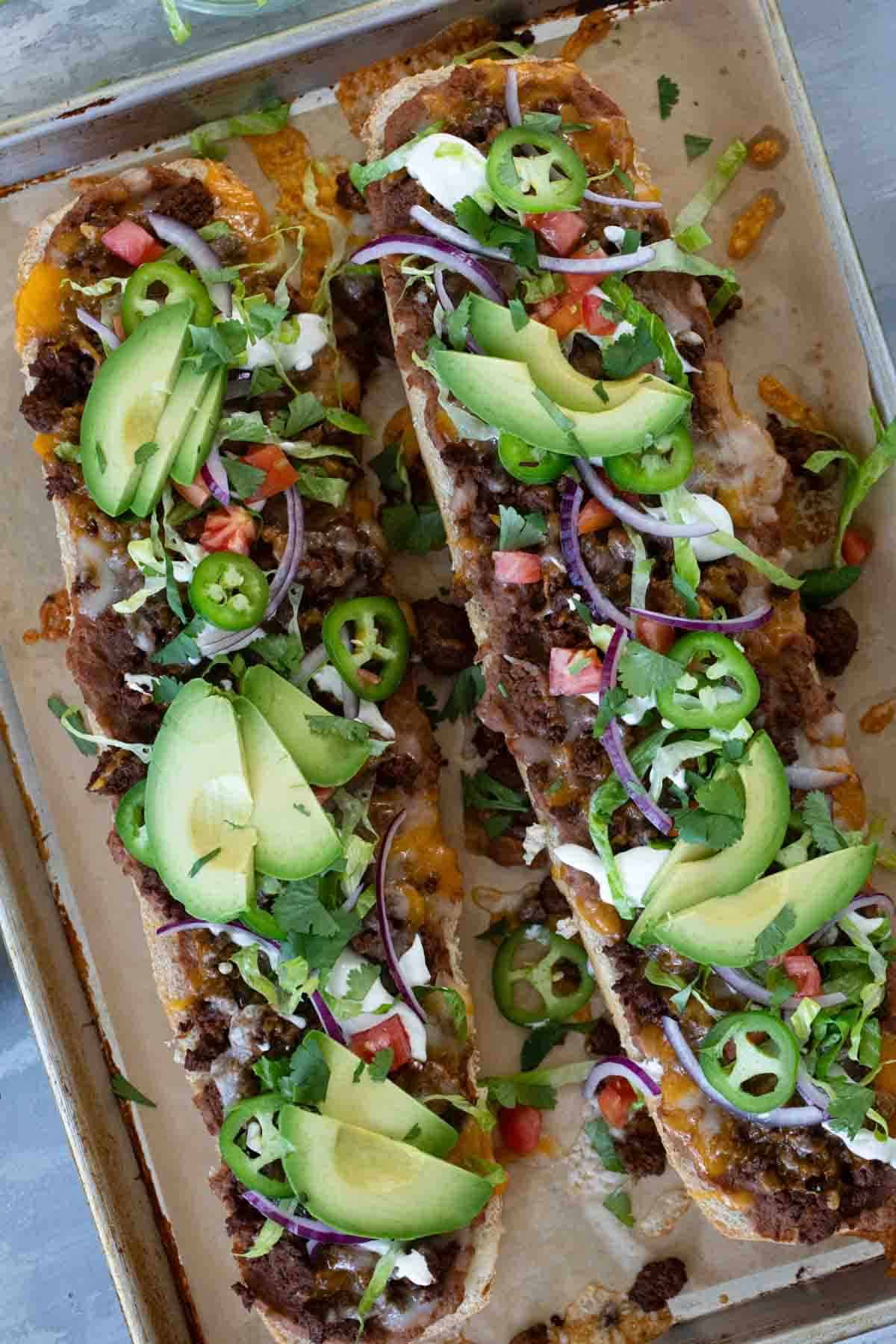 Taco French Bread Pizza on a baking sheet, topped with lettuce, tomatoes, onions, and avocados.